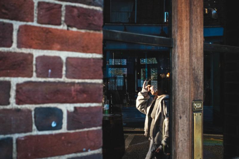 A person is seen reflected in a pub window, with a brick wall covering just left than half of the left of the frame in the foreground.