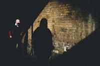 A man in a cap and a silhouetted figure seen in a diagonal shaft of light on a brick wall.