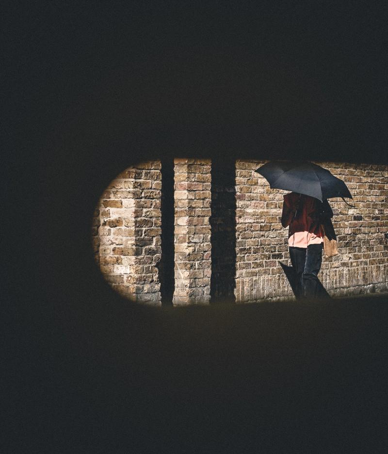 A lady with a parasol is in shadow against a brick wall, seen through a slit in a gate, making two-thirds of the image almost black.