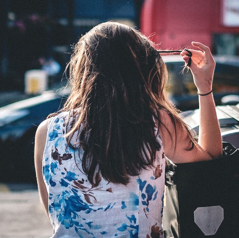 A lady in a white floral dress seen from behind standing on a pavement twiddling her hair.