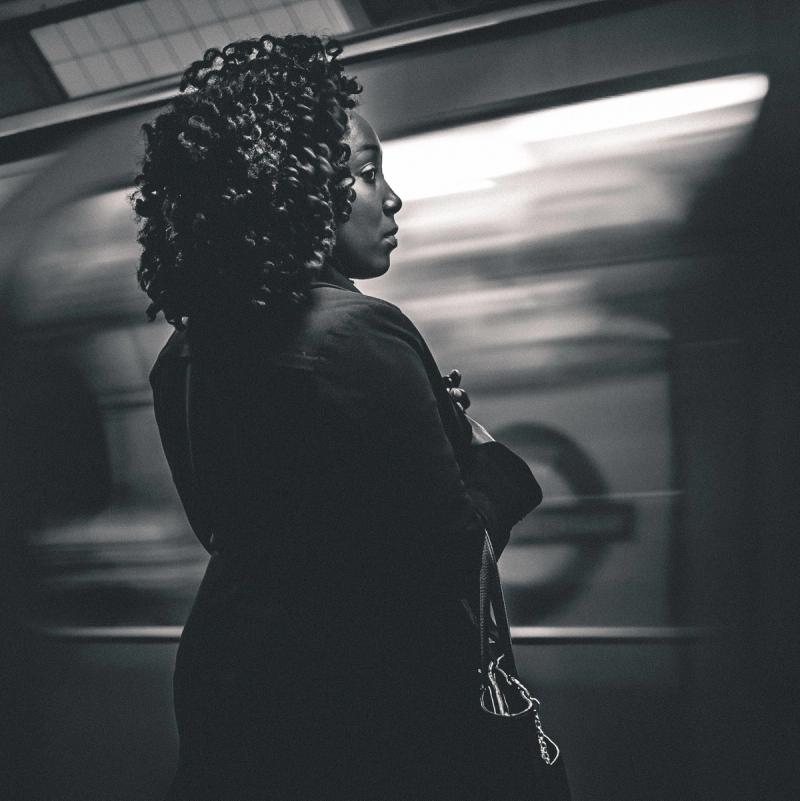 A lady standing on a tube platform seen from behind and to the side. A train is arriving in front of her and it has some motion blur. The underground roundel is partially visible through the window of the train. The image is in black and white.