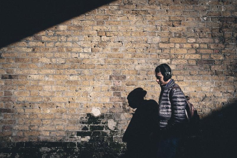 A man in over-the-ear headphones and a puffa-jacket walks through a shaft of diagonal light with his shadow on the brick wall behind.