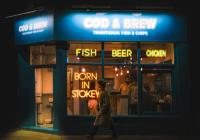 A person walking in front of a hipster-looking chip shop at night. The shop sign, COD & BREW, is illuminated. There are neon signs, FISH, BEER, CHICKEN, and BORN IN STOKEY. 