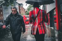 A man in a red tartan jacket and bowler hat carrying a black umbrella walking away from the camera. A man in a black jacket walks towards the camera with a frown and looking down the lens.