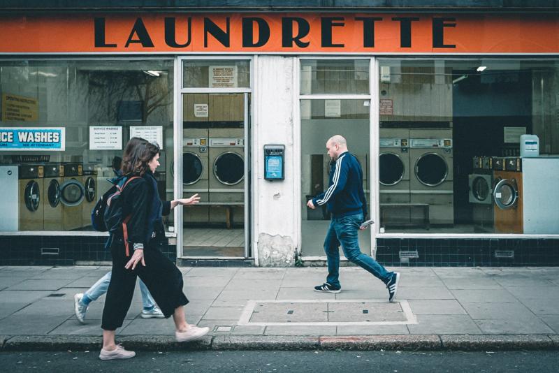 Two ladies walk left-to-right and a man walks right-to-left past a laundrette with an orange sign.