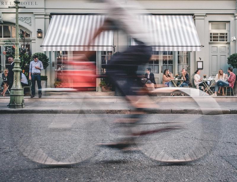 A cyclist is side on in the foreground moving across the frame from the right to the left with some motion blur. An eatery with a striped awning is in the background with some people sitting and standing outside.