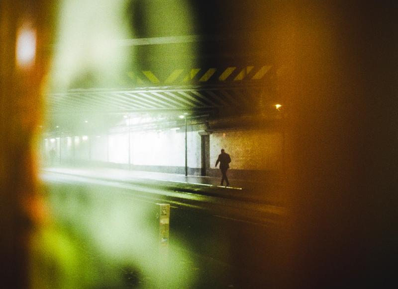 A solitary figure is walking under a railway bridge lit by street lights, framed by a very out-of-focus mirror on the opposite side of the bridge.