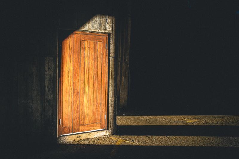 A wooden door in a concrete wall with a ray of sunlight shining onto it across a concrete floor. Most of the rest of the scene is in darkness.