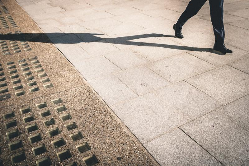 The suited legs and shiny shoes of a man walking up paving stones, with a long shadow of his body trailing behind him.