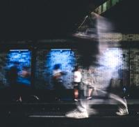 Blurred motion of people walking past a brightly lit brick wall at night.