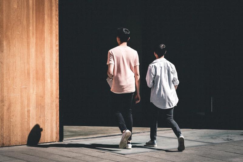 Two people walking away from the camera around a large wooden wall into an area shrouded in darkness.