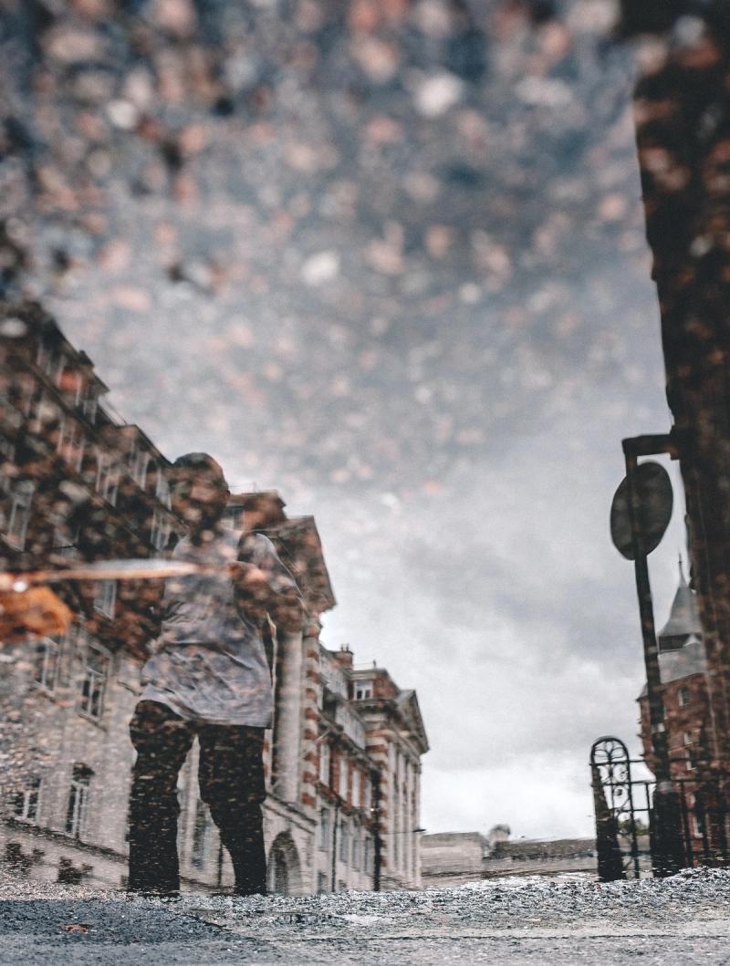 The reflection of a person walking past an old building seen in a puddle.