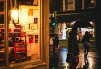 The lit up window of a deli is in the foreground covering the left half of the frame. Three figures are going in different directions in the night rain in the right half of the frame. Two of them have umbrellas. They are slightly reflected in the wet road.