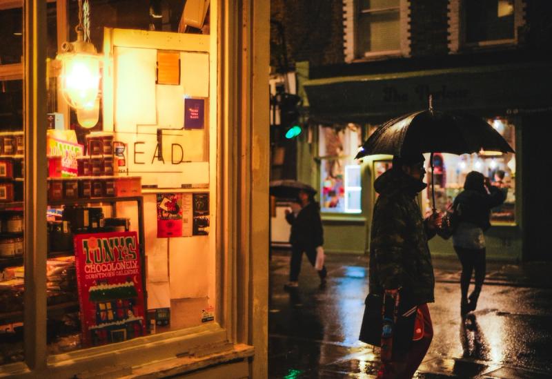 The lit up window of a deli is in the foreground covering the left half of the frame. Three figures are going in different directions in the night rain in the right half of the frame. Two of them have umbrellas. They are slightly reflected in the wet road.