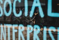 The reflection of a cyclist in a shop window. The window has the words 'SOCIAL ENTERPRISE' written on it.