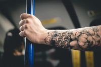 The tattooed arm and hand of a man holding a vertical blue handrail on a London Underground train. The tattoo is a bit like a doodle with some geometric shapes and flowers.