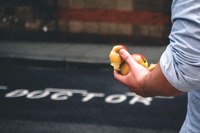 A man's arm and hand holding apples, the word 'DOCTOR' can be seen slightly out of focus on the road behind.