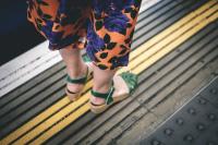 Person wearing green sandals and floral trousers stands next to a yellow safety line on a train platform.