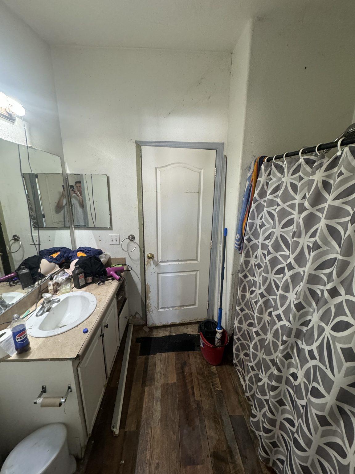 Before the remodel: the original guest bathroom with a single-sink vanity, shower curtain and wood-look floor.