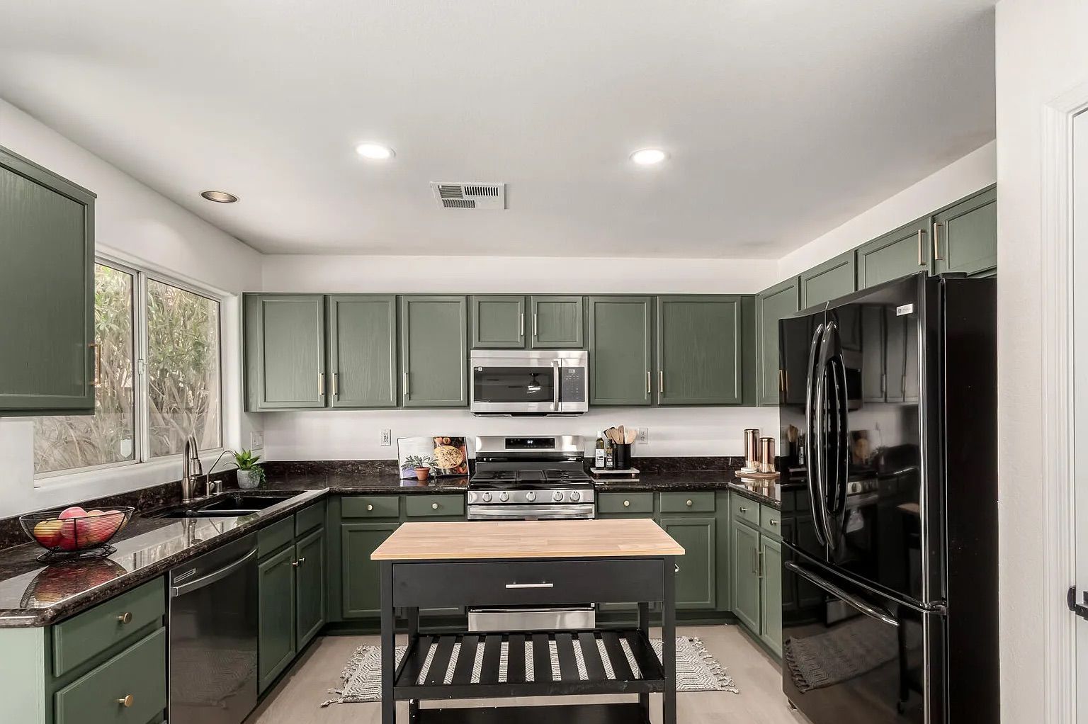 Full view of a kitchen with sage green cabinetry, dark granite countertops, black appliances, and a small rolling prep island in the center.