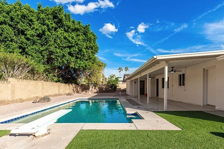 Sunny backyard featuring a large swimming pool with a diving board, artificial turf sections, a long covered patio area, and mature shade trees.