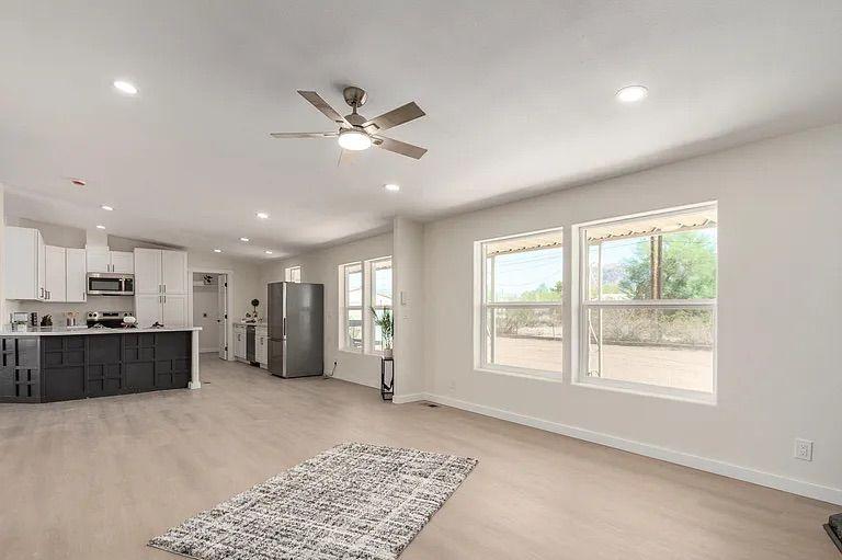 Open concept living and kitchen area featuring light wood flooring, large windows, and a bright kitchen with white cabinets and a dark accent island.