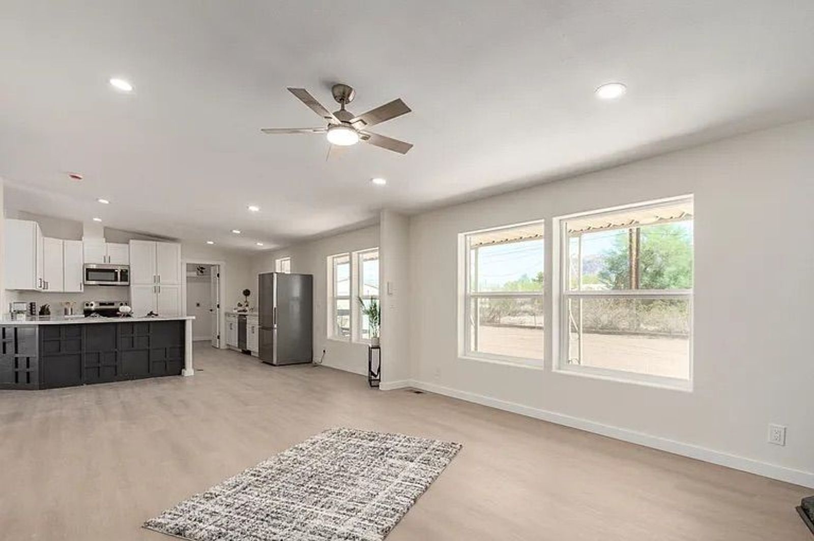 Open concept living and kitchen area featuring light wood flooring, large windows, and a bright kitchen with white cabinets and a dark accent island.