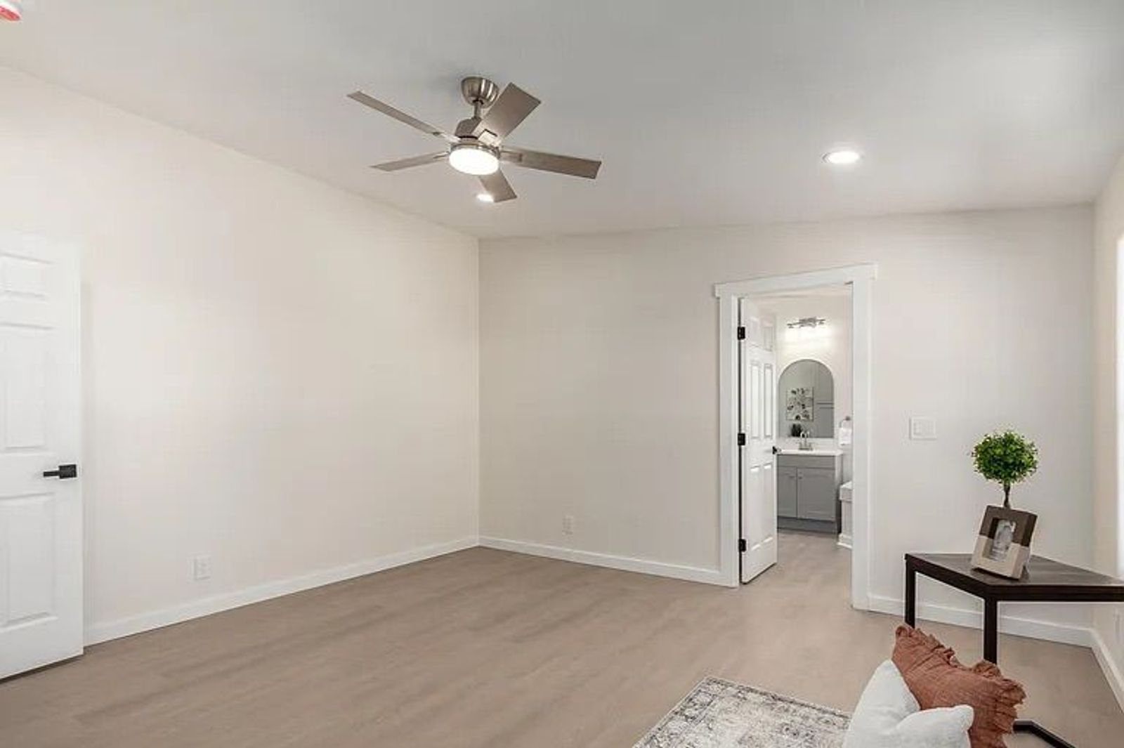 Empty bedroom with light wood flooring, a modern silver ceiling fan, and an open doorway revealing an en-suite bathroom with a grey vanity.