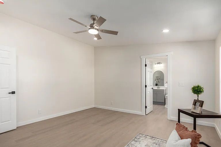 Empty bedroom with light wood flooring, a modern silver ceiling fan, and an open doorway revealing an en-suite bathroom with a grey vanity.