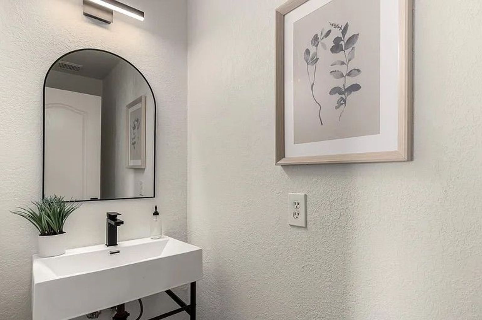 Modern powder room featuring a white trough sink on a black stand, a black-framed arched mirror, and framed botanical wall art.