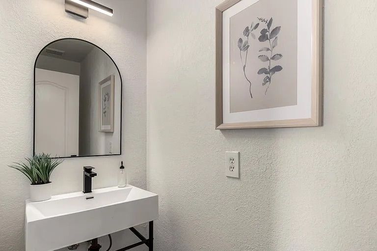 Modern powder room featuring a white trough sink on a black stand, a black-framed arched mirror, and framed botanical wall art.