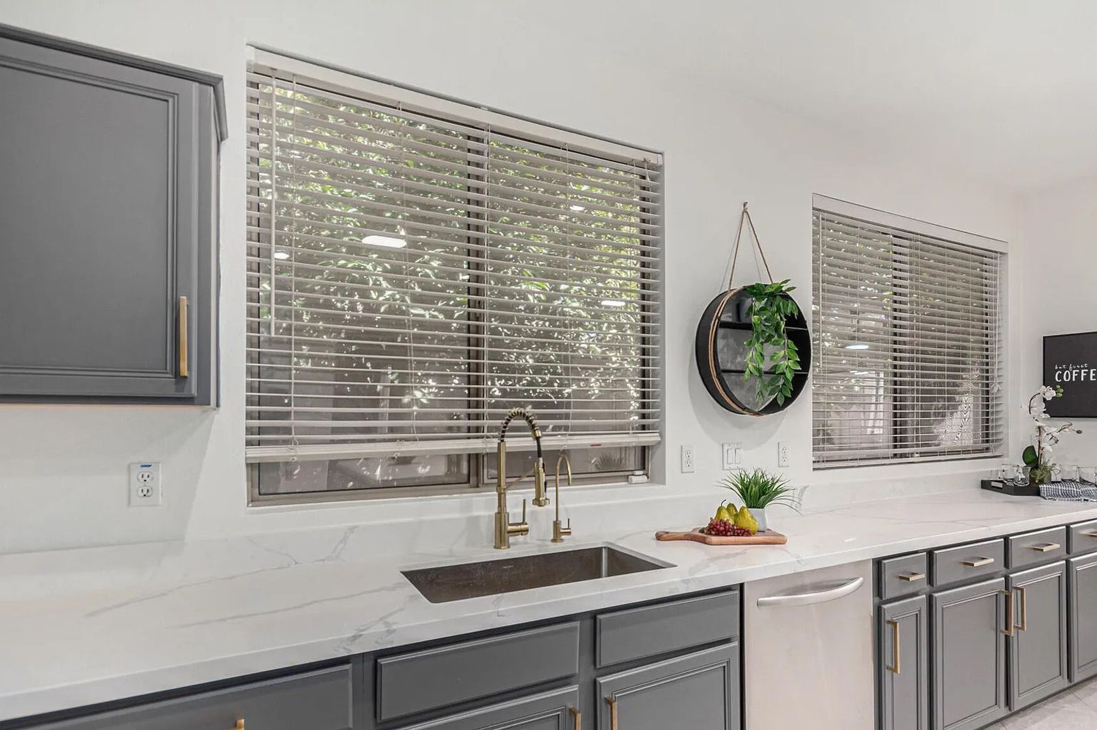 Close-up of a kitchen sink featuring a gold pull-down faucet, undermount stainless steel basin, white quartz countertops, and grey cabinetry.
