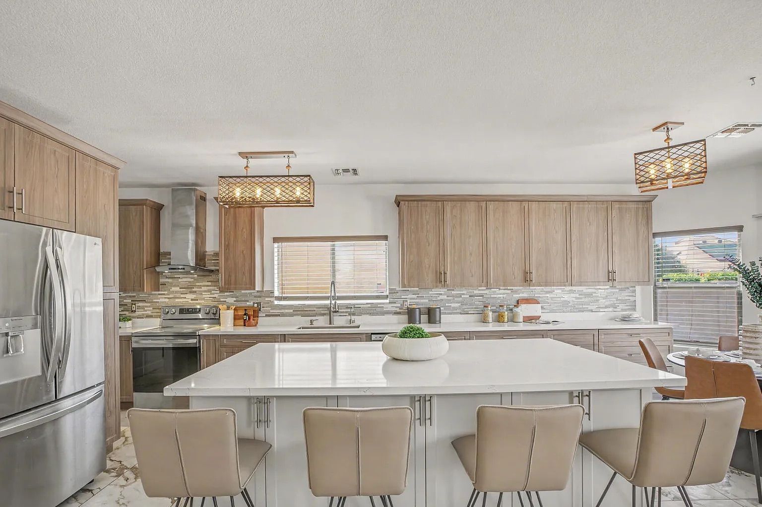 Modern kitchen highlighting a large white quartz island with four beige barstools, light wood-grain cabinetry, stainless steel appliances, and decorative lighting.