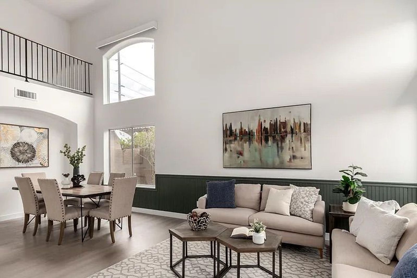 Two-story living room featuring green wainscoting, beige sofas, a dining area, and a second-floor loft balcony overlooking the space.