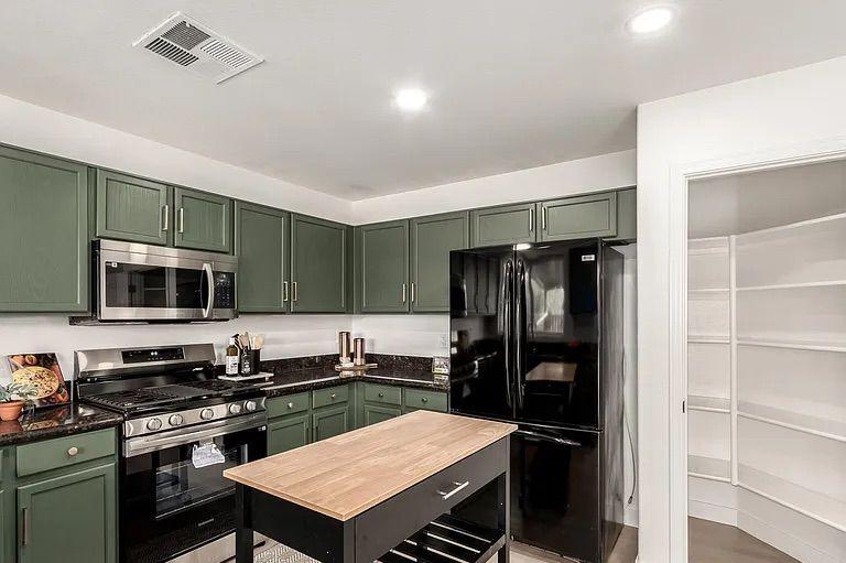 Kitchen area showcasing sage green cabinets, dark countertops, a black refrigerator, a rolling wooden island cart, and an open walk-in pantry.