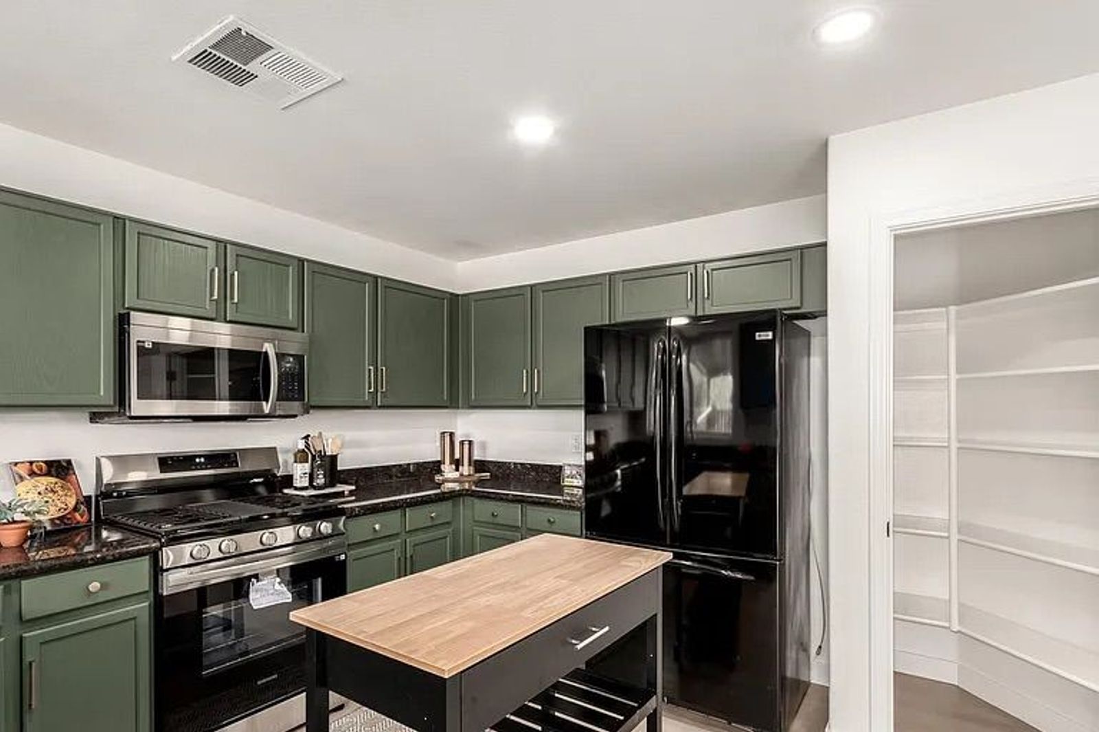 Kitchen area showcasing sage green cabinets, dark countertops, a black refrigerator, a rolling wooden island cart, and an open walk-in pantry.