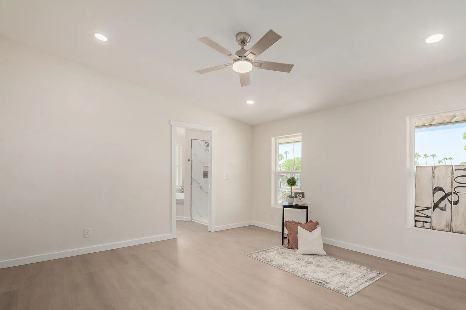 Bright, empty bedroom displaying light wood flooring, a modern ceiling fan, multiple windows, and a doorway leading into an en-suite bathroom.