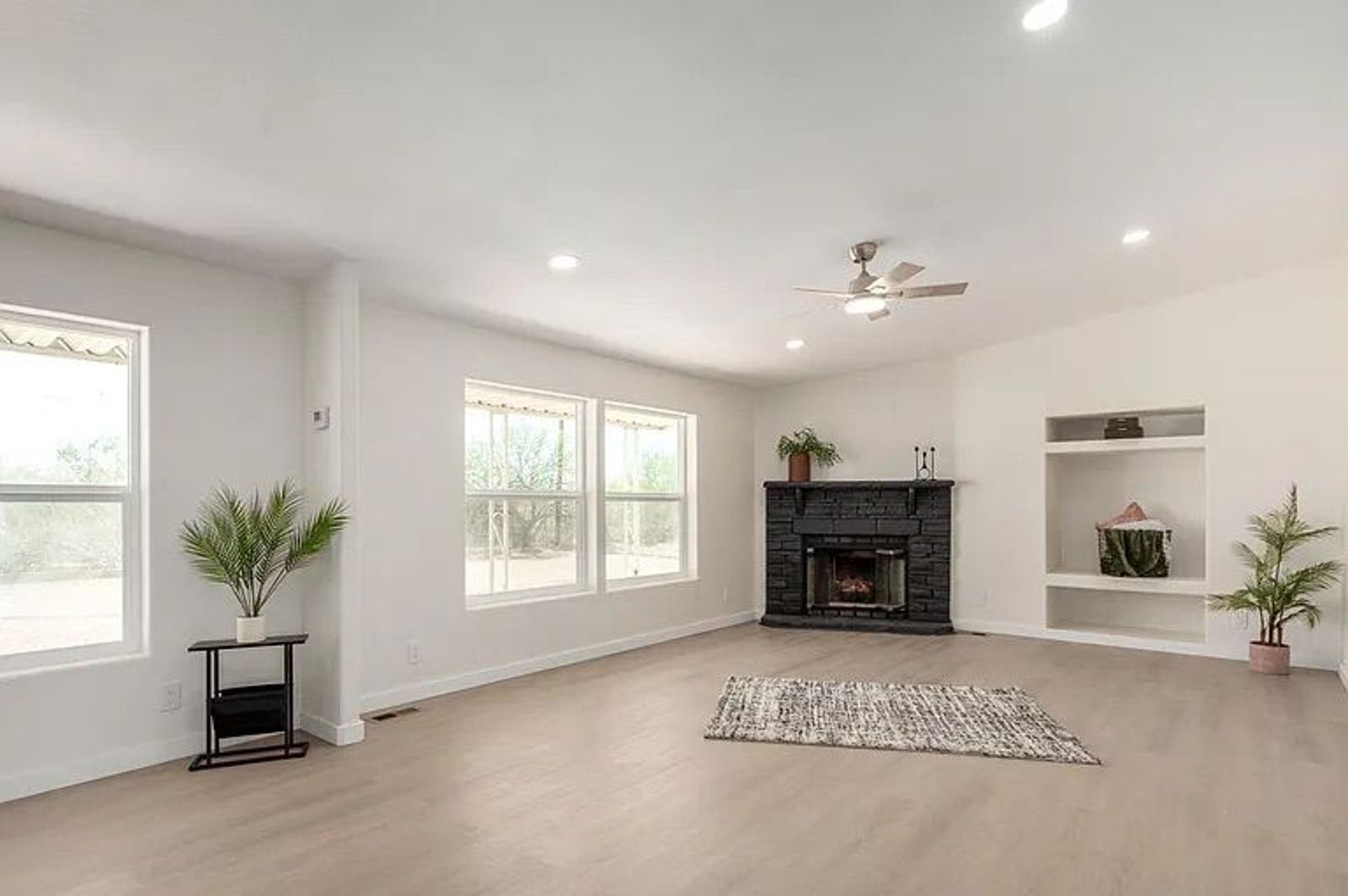 Bright, open living space with large windows, light wood flooring, a black brick corner fireplace, and built-in display shelves.