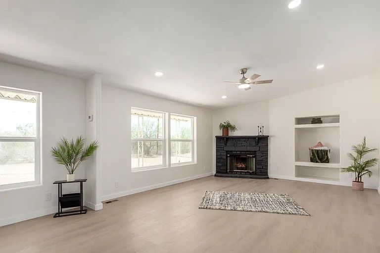Bright, open living space with large windows, light wood flooring, a black brick corner fireplace, and built-in display shelves.