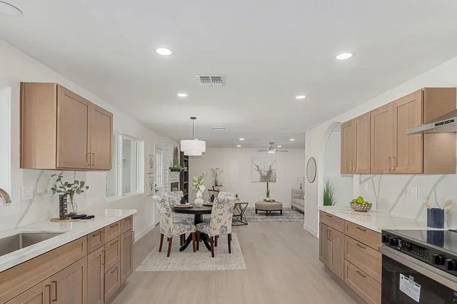 Wide view of an open-concept kitchen and dining area featuring light wood cabinetry, white countertops, stainless steel appliances, and a round dining table leading into a living space.