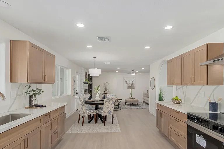 Wide view of an open-concept kitchen and dining area featuring light wood cabinetry, white countertops, stainless steel appliances, and a round dining table leading into a living space.