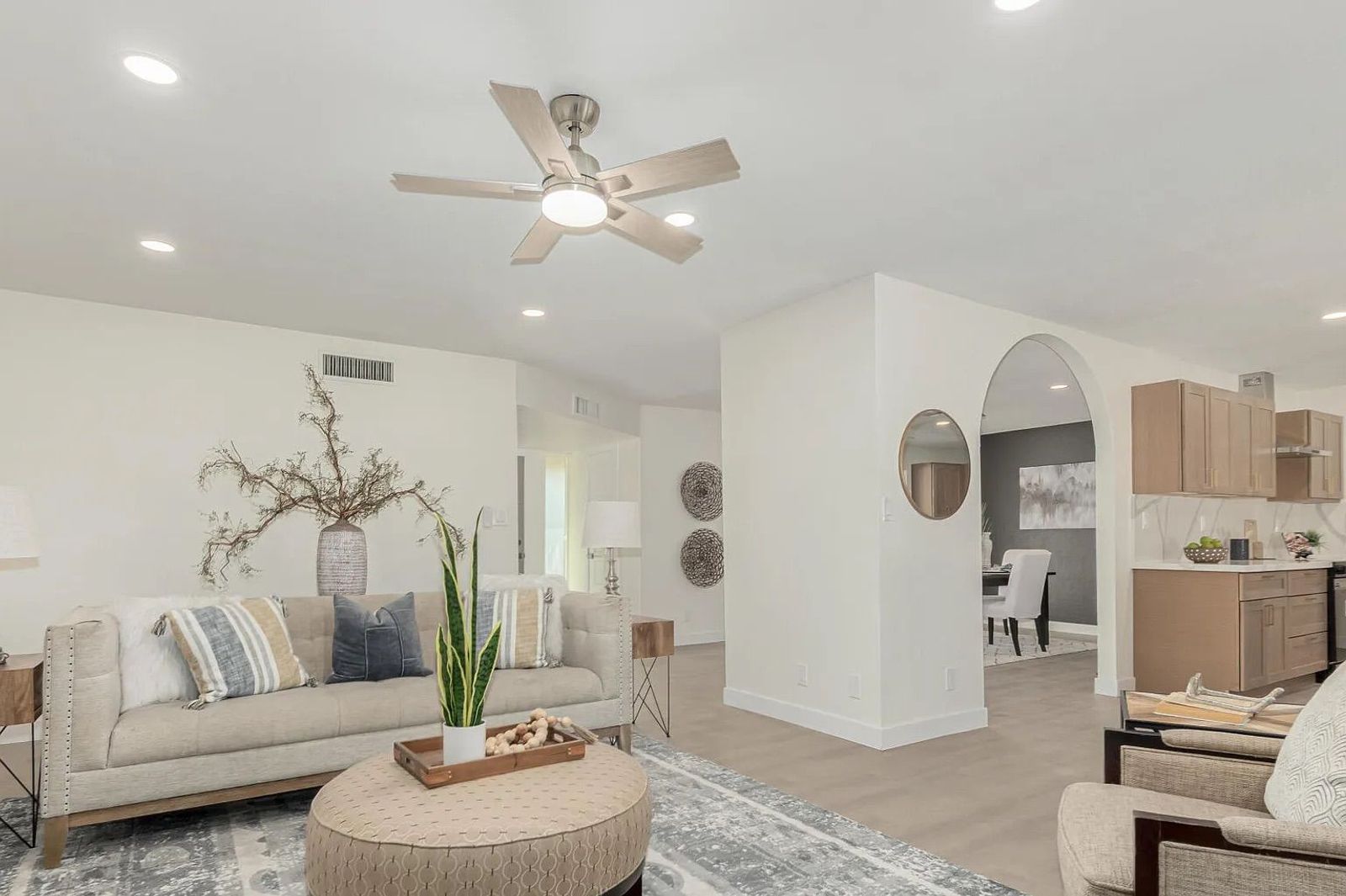 Open-concept living space with a beige sofa, a round tufted ottoman, an arched doorway leading to the dining room, and a view into the kitchen.