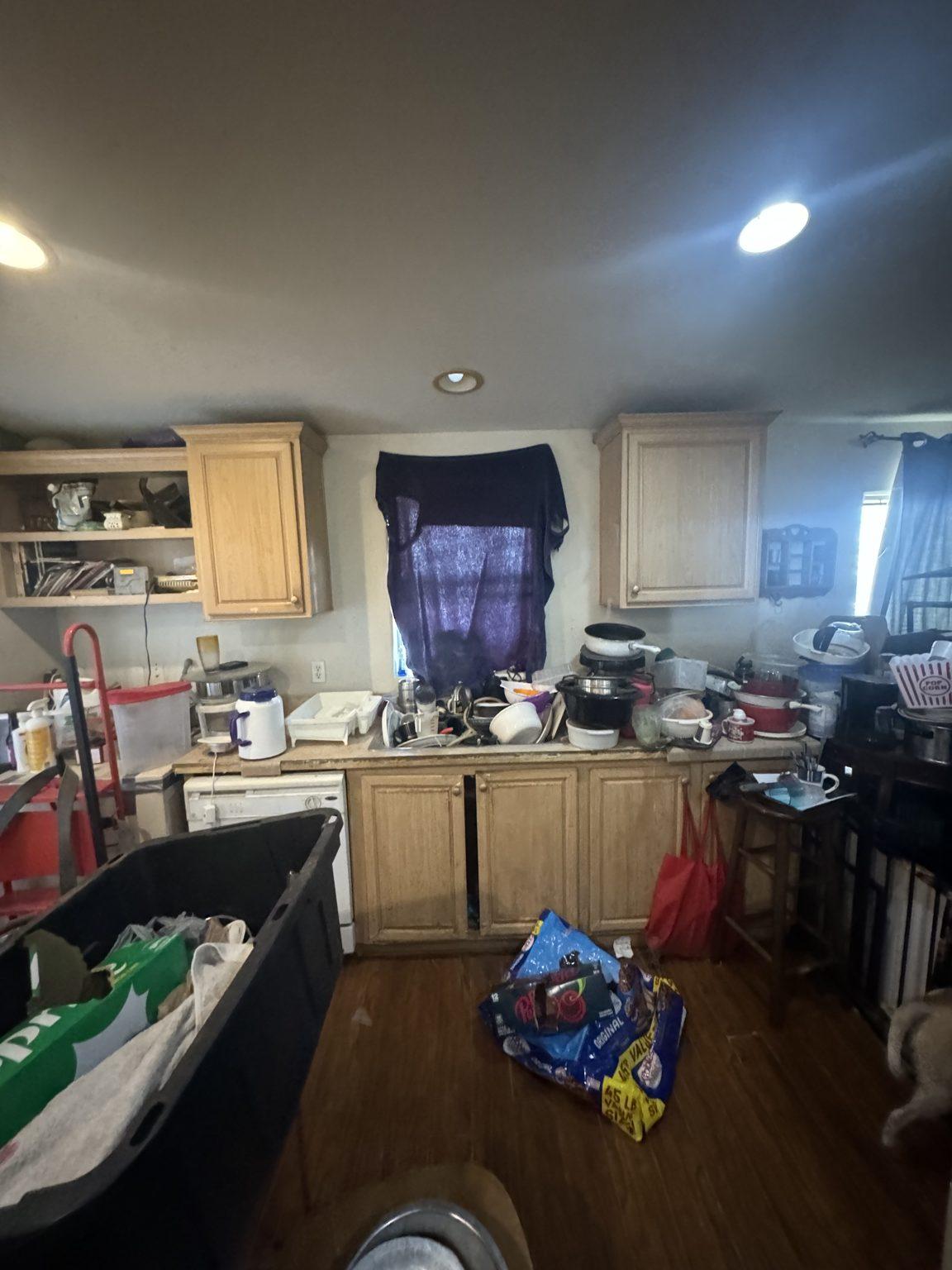 Before the remodel: the original kitchen sink wall with oak cabinets, a curtained window and white dishwasher.