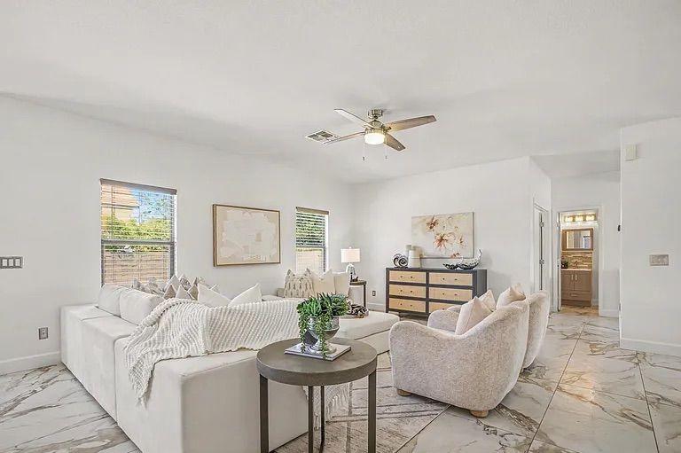 Bright living room displaying a large white sectional sofa, two textured armchairs, a round accent table, and marble-look tile floors, with a view down a hallway.