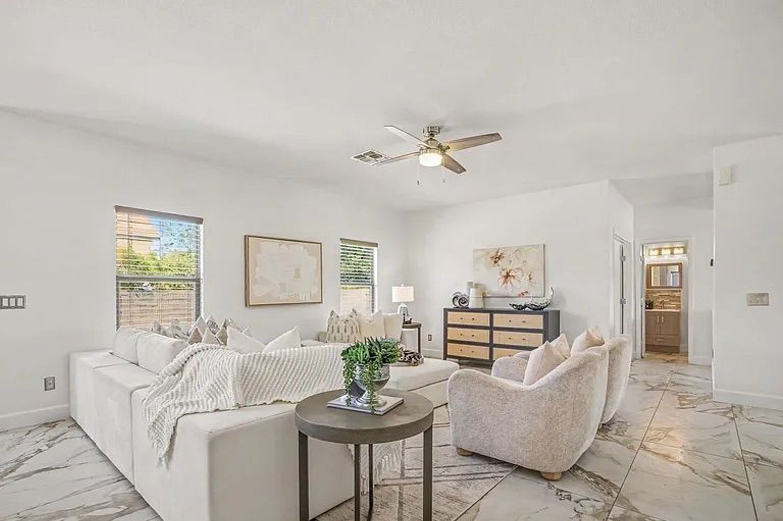 Bright living room displaying a large white sectional sofa, two textured armchairs, a round accent table, and marble-look tile floors, with a view down a hallway.