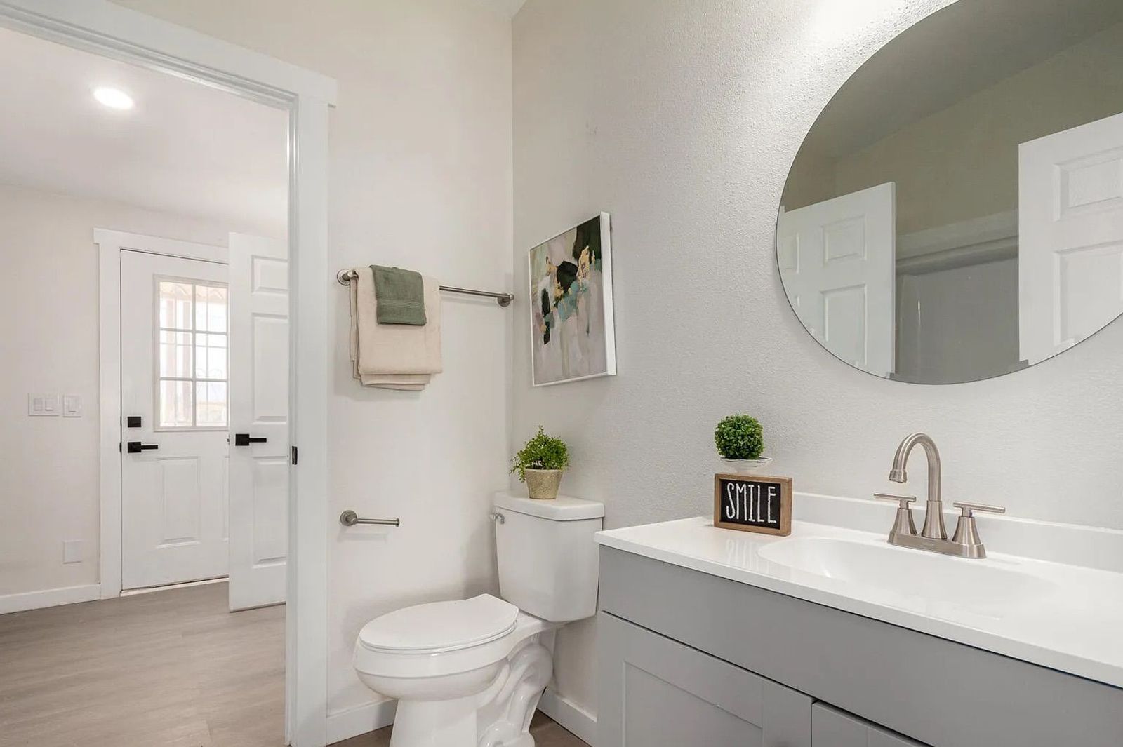 Bathroom showcasing a grey vanity with a round wall mirror, a white toilet, light wood flooring, and an open doorway leading to a back entryway.