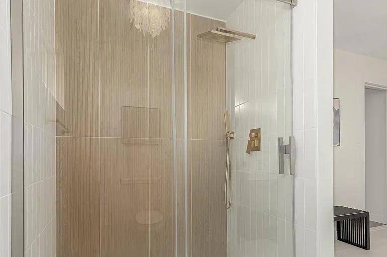 Modern glass-enclosed shower featuring a vertical wood-look accent tile wall, white tiled side walls, and brushed gold fixtures including a square rain showerhead.