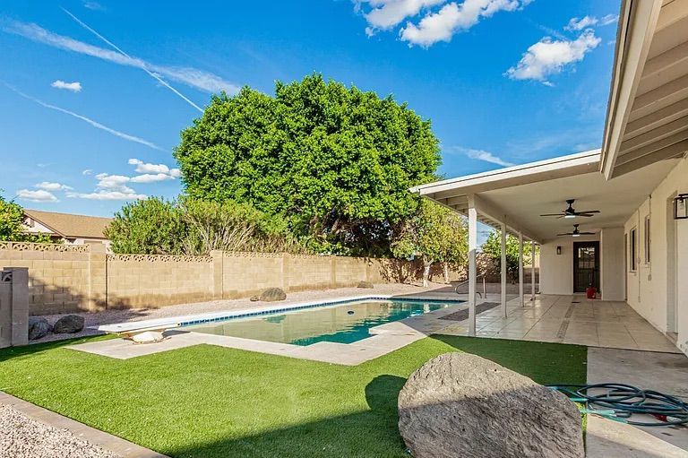 Large backyard space featuring a swimming pool with a diving board, patches of artificial turf, a long covered patio, and a mature green shade tree.