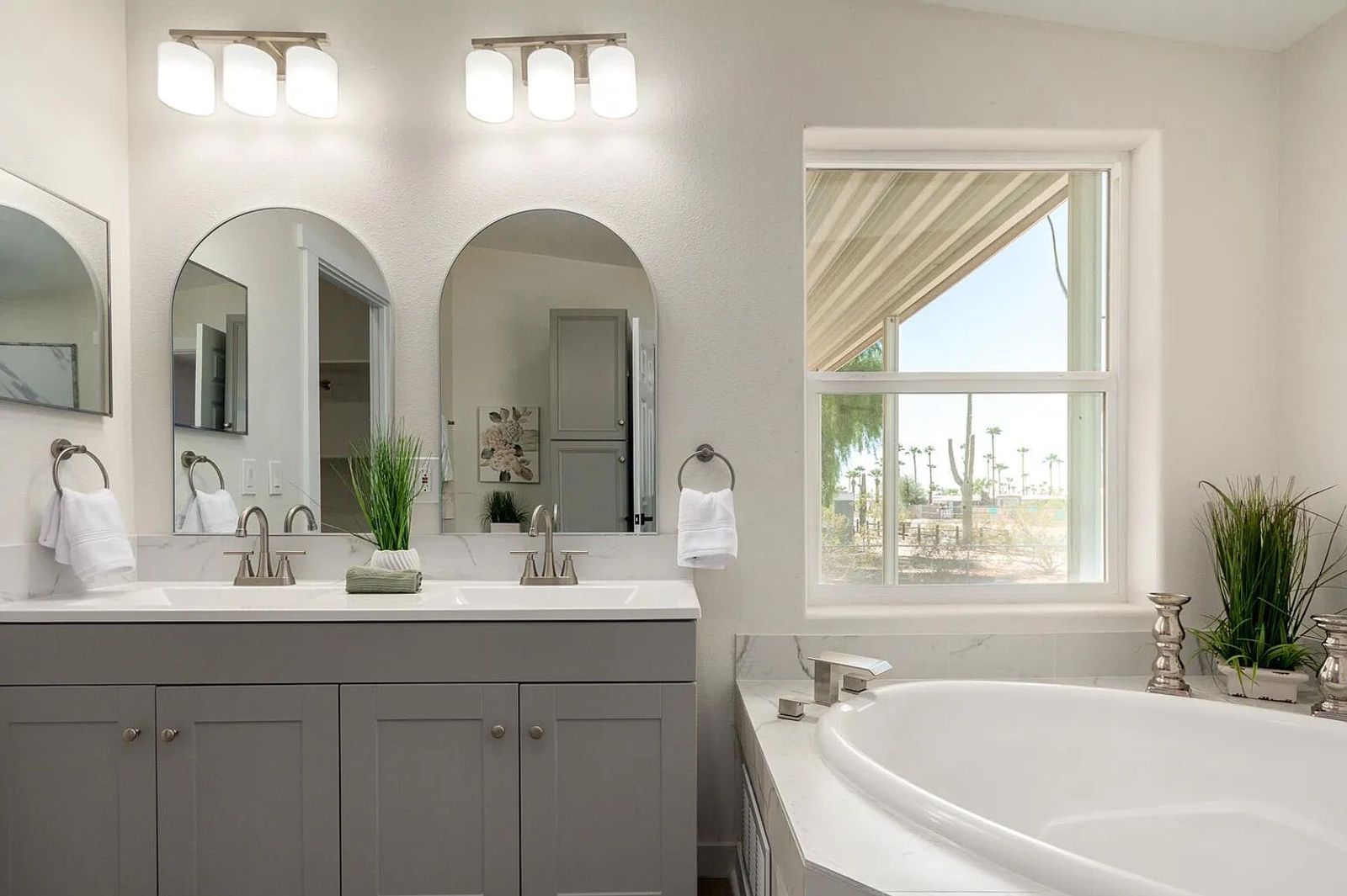 Close-up of a bathroom vanity area showing dual sinks, brushed nickel hardware, grey cabinetry, arched mirrors, and a corner bathtub beside a window.