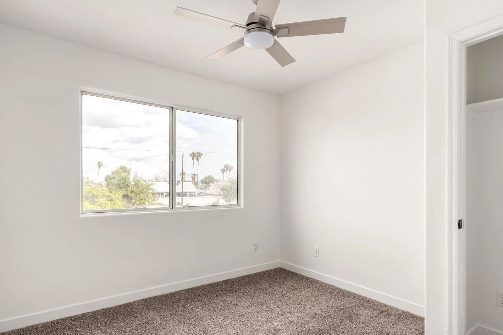 Empty bedroom space with tan carpeting, white walls, a ceiling fan, a view from the window, and an open closet space.